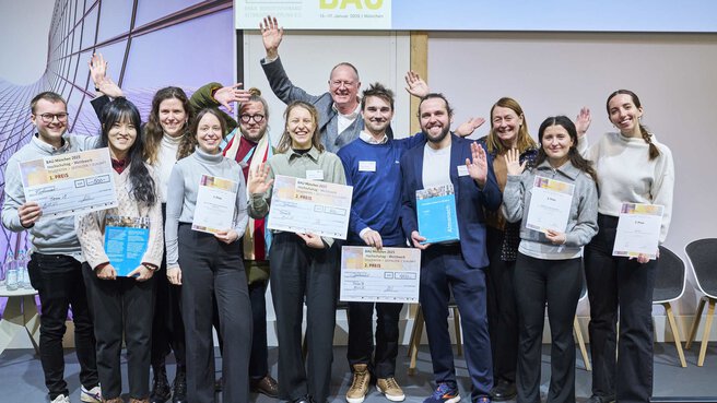 A group of twelve people with winning certificates wave smilingly for a group photo.