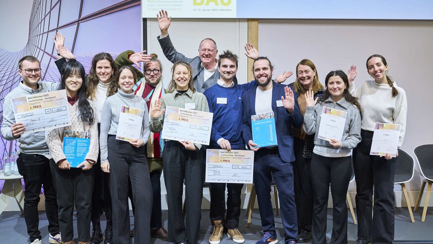 Eine Gruppe von zwölf Personen mit Siegerurkunden winkt lächelnd für ein Gruppenfoto.