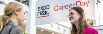 Two women are holding leaflets and chatting. Above them hangs a sign featuring the EXPO REAL logo and the red text ‘CareerDay’.