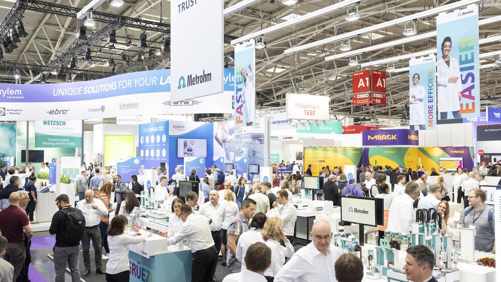 A busy trade fair with lots of people visiting stands and exhibitions in a large, brightly lit exhibition hall.