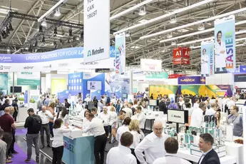 A busy trade fair with lots of people visiting stands and exhibitions in a large, brightly lit exhibition hall.