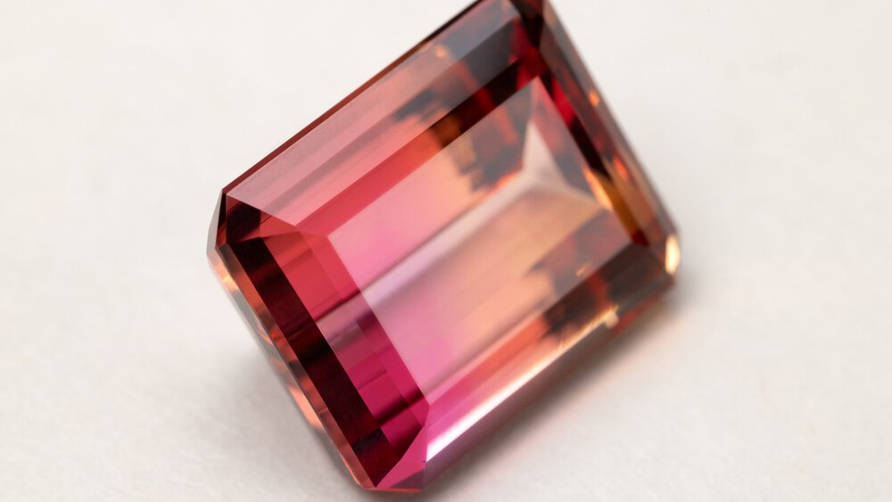 The close-up view of a rectangular cut gemstone in several shades of red resting on a white surface.