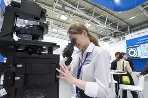 A woman in a lab coat looks through a large electronic microscope at an exhibition stand