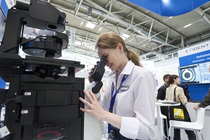 A woman in a lab coat looks through a large electronic microscope at an exhibition stand