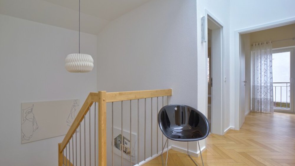 Minimalistischer Flur mit rundem schwarzen Stuhl, Holzgeländer und weißer Hängelampe über der Treppe.