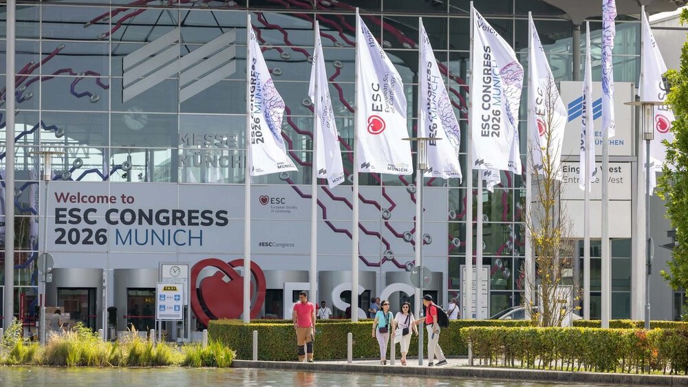 Vor einem modernen Glasgebäude, das sich in einem Teich spiegelt, gehen Menschen an Fahnen und einem großen Eingangsschild mit der Aufschrift „Willkommen beim ESC-Kongress 2026 in München“ vorbei.
