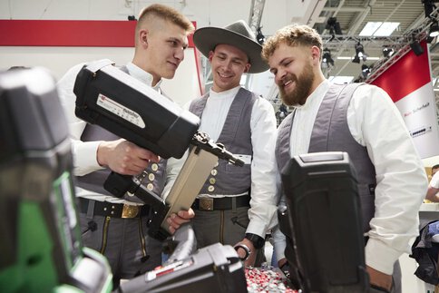 Drei Männer in grauer Kluft untersuchen lächelnd ein großes Bolzenschubgerät an einem Messestand.