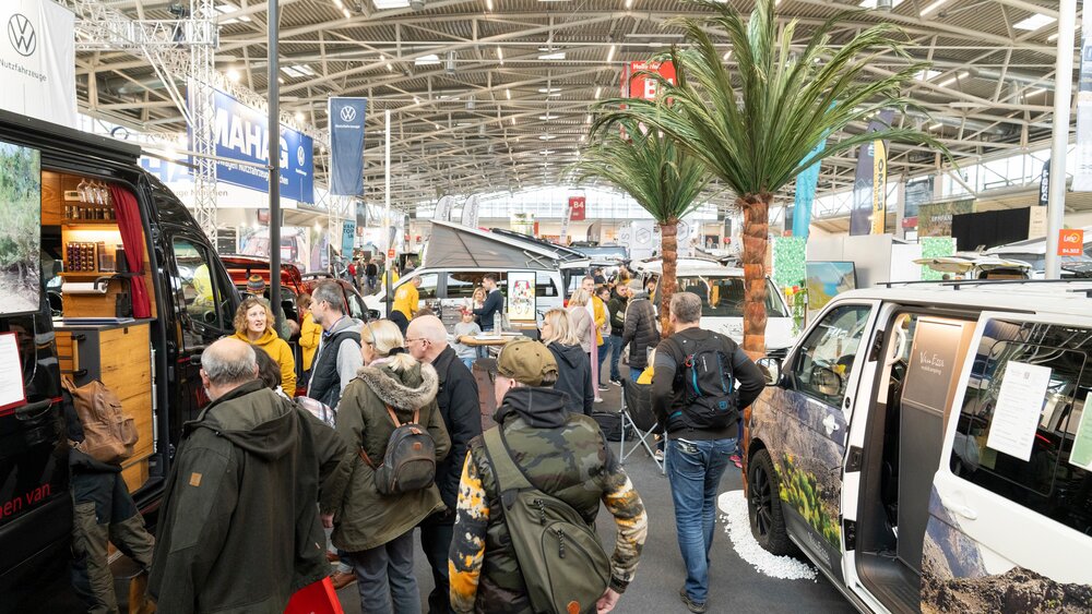 Besucherinnen und Besucher informieren sich in einer Messehalle zwischen ausgestellten Campervans und Reisemobilen über Angebote rund um mobiles Reisen.