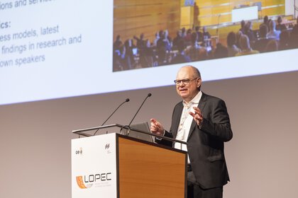 A man in a suit speaks on a podium with LOPEC branding before a presentation.