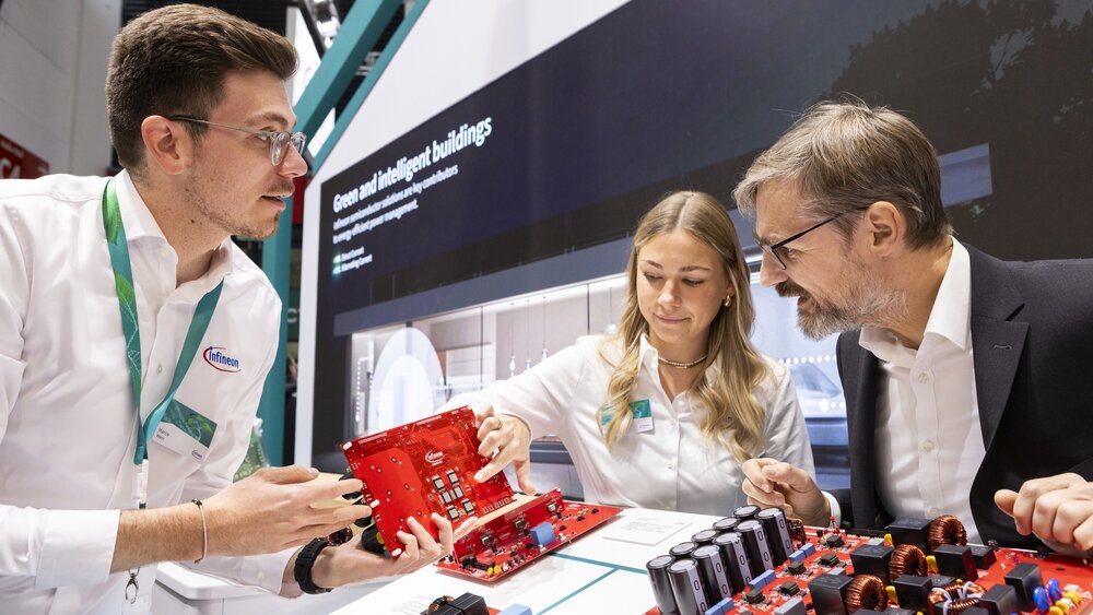 Ein Mann und eine Frau in weißen Oberhemden erklären einem interessierten Herrn im Anzug eine von mehreren unterschiedlichen roten Leiterplatinen.