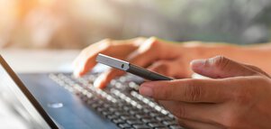 Hands type on a laptop keyboard while holding a smartphone.