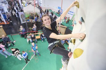 Eine Frau klettert an einer Indoor-Kletterwand und lächelt in die Kamera. Sie wird unten von einer Person gesichert.