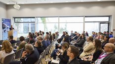 A conference room with rows of seated people looking at a speaker in front, standing behind a podium with a laptop