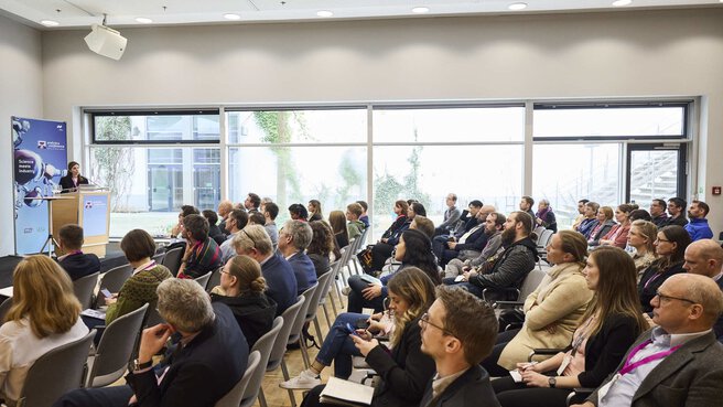 Ein Konferenzraum mit Reihen von sitzenden Personen, die auf einen Redner vorne schauen, der hinter einem Podium steht mit einem Laptop