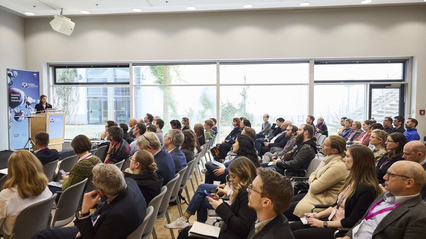 A conference room with rows of seated people looking at a speaker in front, standing behind a podium with a laptop