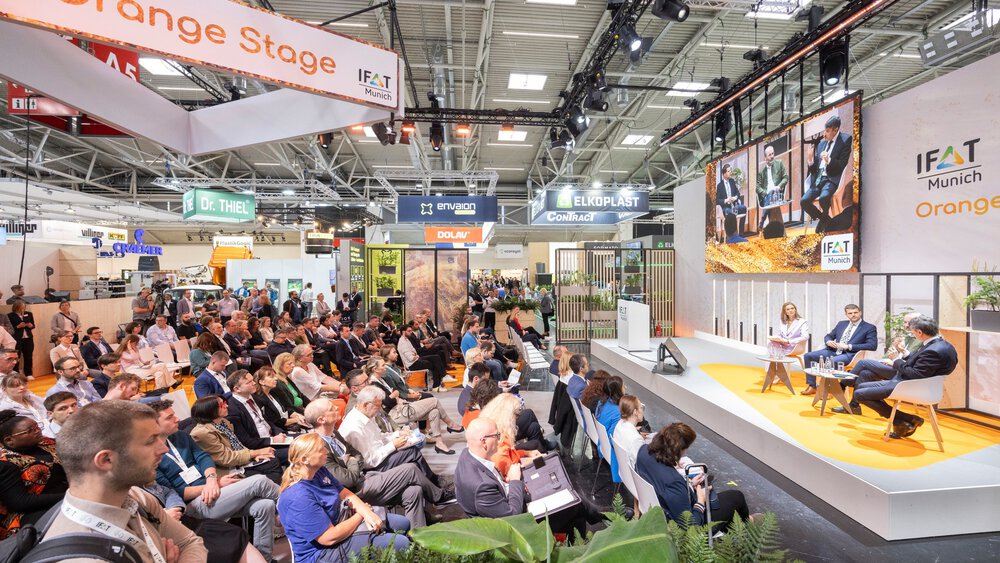 Audience attending a panel discussion on the Orange Stage at IFAT Munich inside an exhibition hall.