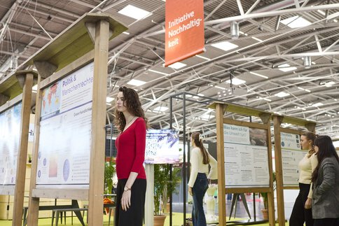 Besucherinnen der Messe f.re.e betrachten Infotafeln zur Nachhaltigkeit, umgeben von Holzaufstellern und einem Banner zur Initiative.