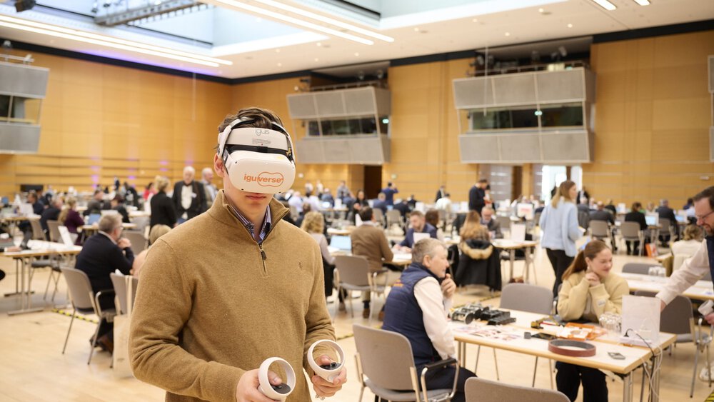 A young man wearing an Iguverse VR headset holds two VR controllers in his hands. In the background, several participants of the bauma Mediendialog 2025 are seated at tables, engaged in discussions and presentations.