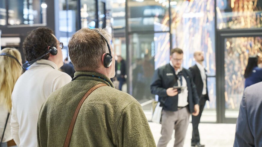 Mehrere Besucher der BAU stehen mit Kopfhörern in einer Ausstellungshalle und blicken nach rechts.