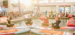 Vier Personen machen Yoga auf Paddleboards in einem Wasserbecken auf einer Messe, umgeben von Ständen und Besuchern.