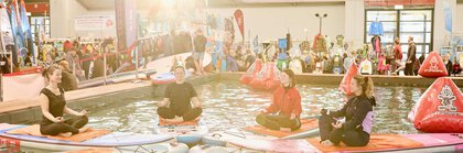Vier Personen machen Yoga auf Paddleboards in einem Wasserbecken auf einer Messe, umgeben von Ständen und Besuchern.