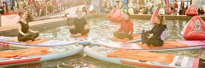 Vier Personen machen Yoga auf Paddleboards in einem Wasserbecken auf einer Messe, umgeben von Ständen und Besuchern.
