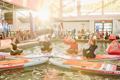Vier Personen machen Yoga auf Paddleboards in einem Wasserbecken auf einer Messe, umgeben von Ständen und Besuchern.