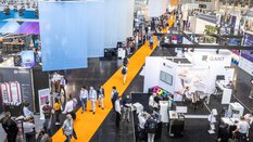 A busy exhibition hall with people walking across a bright yellow carpet, with various stands in the background.