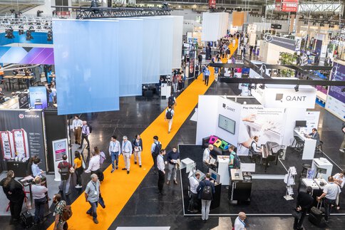 A busy exhibition hall with people walking across a bright yellow carpet, with various stands in the background.