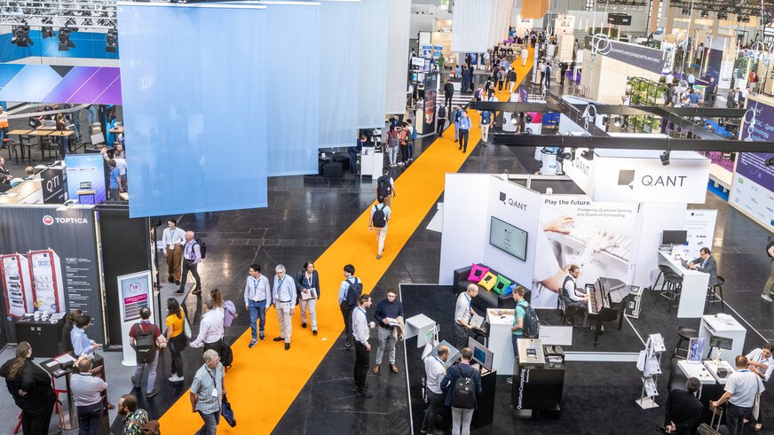 A busy exhibition hall with people walking across a bright yellow carpet, with various stands in the background.