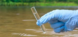 Eine Hand in einem blauen Kunststoff-Handschuh hält eine Reagenzglasprobe mit Wasser aus einem Fluss.