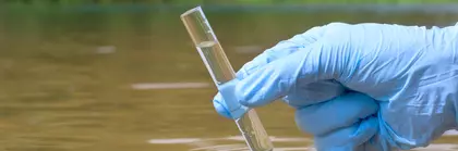 A hand in a blue plastic glove holds a test tube sample with water from a river.
