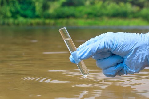 A hand in a blue plastic glove holds a test tube sample with water from a river.