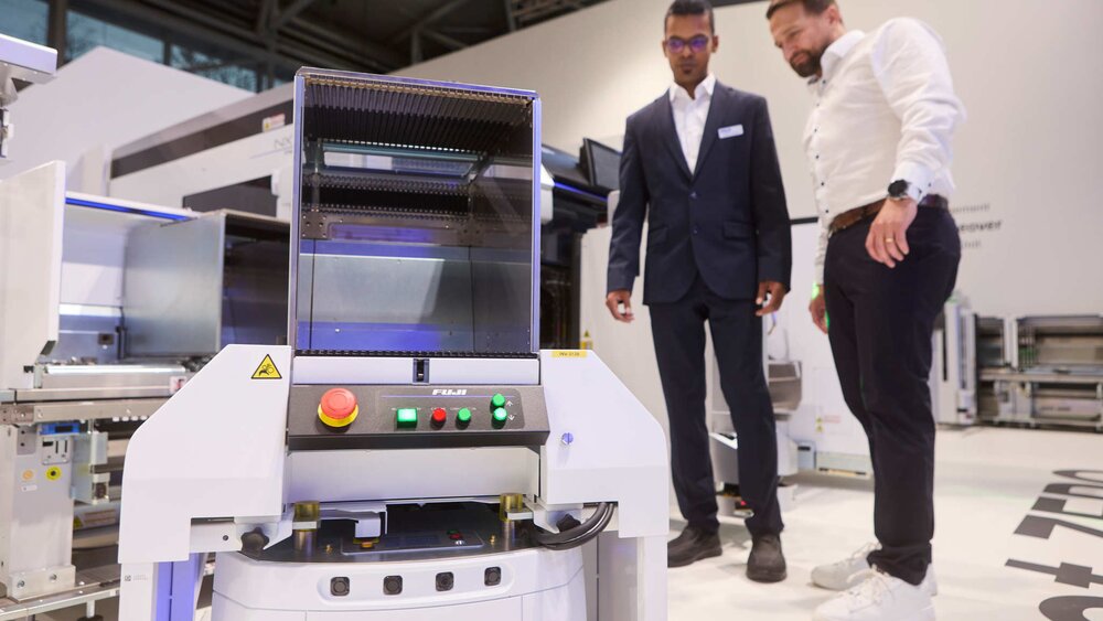 Zwei Männer in Anzügen betrachten eine weiße Maschine mit der Bezeichnung „OMRON“, die aus einem Bedienfeld, Kabeln und einem aufgesetzten Kubus besteht.