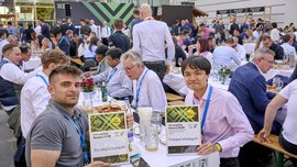 Two winners of the 2025 Innovation Award present their certificates. They are seated at tables set with place settings and beer benches. Many people can be seen in the background.