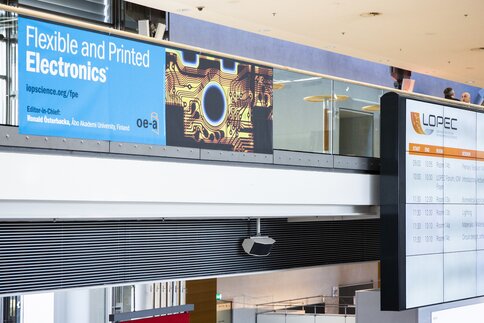 Close-up of an OE-A advertising banner with the text ‘Flexible and Printed Electronics’ and next to it a large screen showing LOPEC trade fair events.