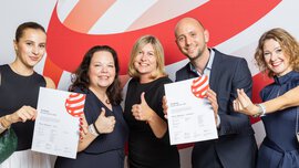 Fünf Menschen lächeln, halten Urkunden in der Hand und zeigen den Daumen nach oben vor einem rot-weißen Hintergrund.