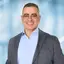 Ein lächelnder Mann mit eckiger Brille, grauem Jackett und blauem Hemd.