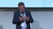 A man in a suit gives a presentation in front of a projector and gestures with his hands.