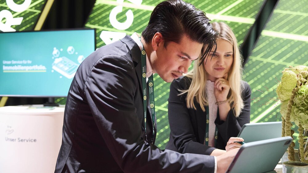 Zwei junge Fachleute im Anzug arbeiten gemeinsam an Tablets an einem Messestand der EXPO REAL. Im Hintergrund ist ein grüner Bildschirm mit digitalen Grafiken und dem Schriftzug „Unser Service für Ihr Immobilienportfolio“ zu sehen. Die Szene zeigt konzentrierte Zusammenarbeit in einem modernen, technologieorientierten Umfeld.