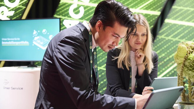 Zwei junge Fachleute im Anzug arbeiten gemeinsam an Tablets an einem Messestand der EXPO REAL. Im Hintergrund ist ein grüner Bildschirm mit digitalen Grafiken und dem Schriftzug „Unser Service für Ihr Immobilienportfolio“ zu sehen. Die Szene zeigt konzentrierte Zusammenarbeit in einem modernen, technologieorientierten Umfeld.