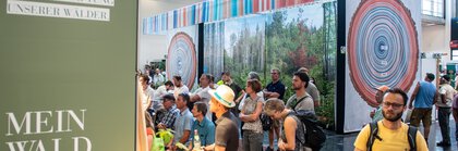 Die Besucher versammeln sich bei einer Ausstellung in den Innenräumen, die mit Exponaten zum Thema Wald, Informationstafeln und einer großen Kulisse mit Baumringen und einer Waldszene gestaltet ist.