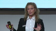 Eine Frau im Blazer spricht ins Headset-Mikrofon und hält eine Fernbedienung vor einem Bildschirm.