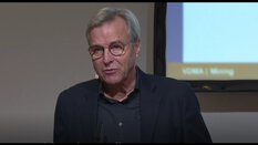 Ein grauhaariger Mann mit Brille spricht vor einem Bildschirm während einer Präsentation.