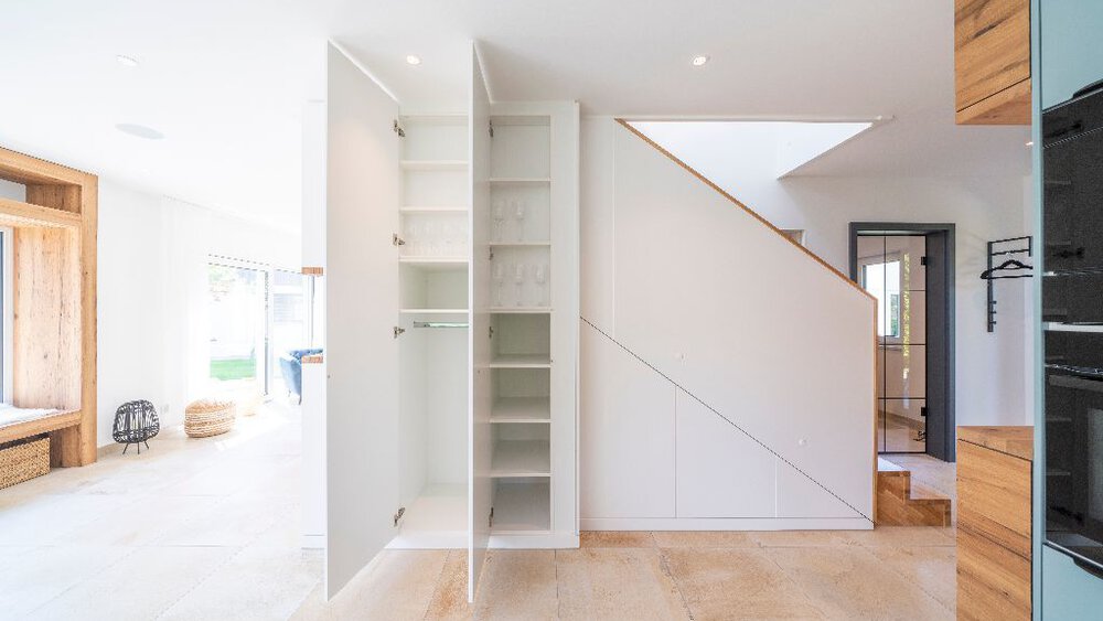 Minimalistisches Interieur mit weißen Wänden, Holzboden, offenen Regalen und einer Treppe im Hintergrund.