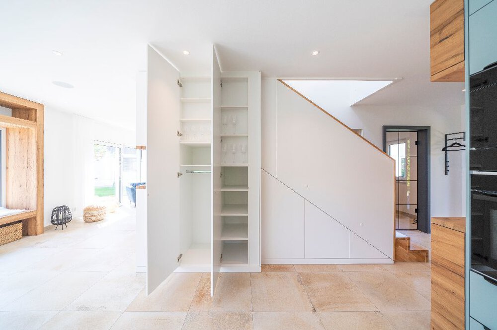 Minimalistisches Interieur mit weißen Wänden, Holzboden, offenen Regalen und einer Treppe im Hintergrund.