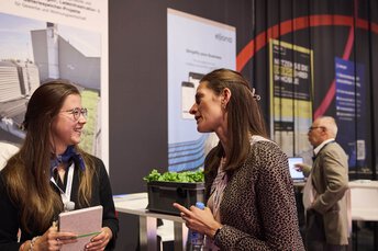 Zwei Frauen im Messeumfeld führen ein freundliches Gespräch, eine hält ein Notizbuch und die andere eine Wasserflasche. Im Hintergrund sind weitere Besucher sowie Informationsposter und ein Messestand zu sehen.