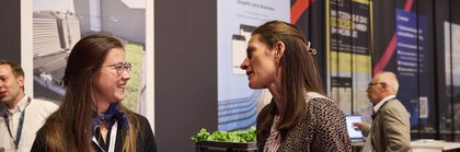 Zwei Frauen im Messeumfeld führen ein freundliches Gespräch, eine hält ein Notizbuch und die andere eine Wasserflasche. Im Hintergrund sind weitere Besucher sowie Informationsposter und ein Messestand zu sehen.
