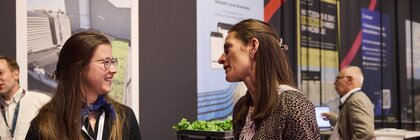 Zwei Frauen im Messeumfeld führen ein freundliches Gespräch, eine hält ein Notizbuch und die andere eine Wasserflasche. Im Hintergrund sind weitere Besucher sowie Informationsposter und ein Messestand zu sehen.