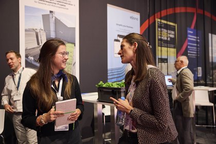 Zwei Frauen im Messeumfeld führen ein freundliches Gespräch, eine hält ein Notizbuch und die andere eine Wasserflasche. Im Hintergrund sind weitere Besucher sowie Informationsposter und ein Messestand zu sehen.